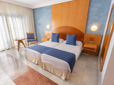 Helles Schlafzimmer mit Doppelbett, blauen Kissen, Holzkopfteil, Nachttischen und Fenster mit Gardinen.