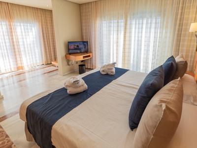 Helles Schlafzimmer mit großem Bett, dunkelblauer Tagesdecke, zwei Handtüchern und TV an der Wand.