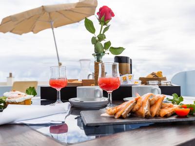 Tisch mit Drinks, Ebly-Hühnchen, roter Rose und Sonnenschirm auf Terrasse bei bewölktem Himmel.