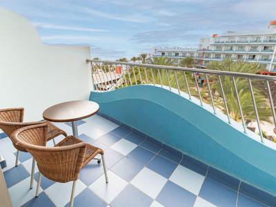 Balkon mit blauer Brüstung, weiß-blau kariertem Boden, zwei runden Tischen und braunen Stühlen.