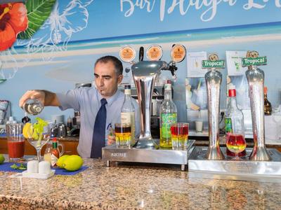 Mann schenkt Bier an Bar mit bunten Getränken, Zapfanlagen und tropischer Dekoration ein.