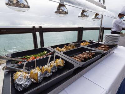 Grillstation mit gegrilltem Fleisch, Gemüse und Brot auf einer Terrasse mit Meerblick bei bewölktem Wetter.