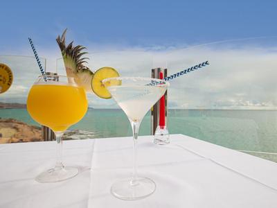 Zwei bunte Cocktails auf weiß gedecktem Tisch mit Meeresblick und blauem Himmel im Hintergrund.