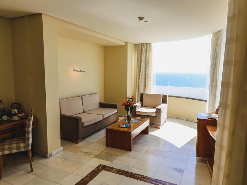 Camera d'albergo luminosa con vista mare, divani, poltrone e scrivania.