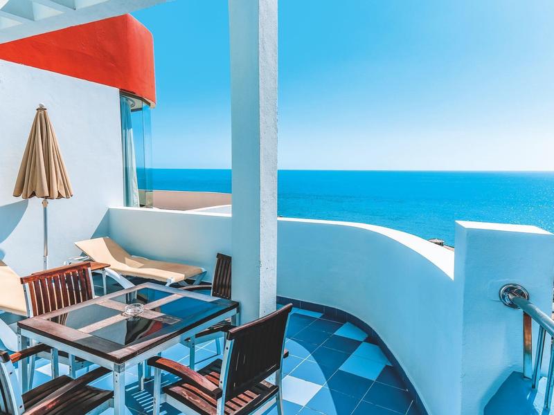 Balcone con vista sul mare blu, tavolo e sedie, ombrellone accanto a muro bianco.