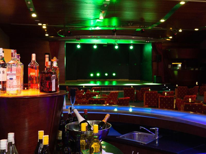 Un bar elegante con bancone illuminato, bottiglie e sedie in una stanza buia con illuminazione verde sullo sfondo.