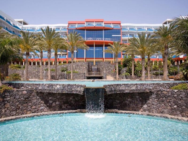 Hotel moderno con palme e cascata davanti a una piscina limpida.