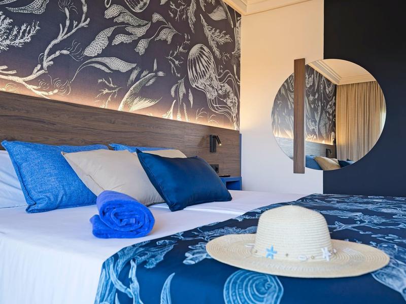 Camera d'hotel luminosa con letto blu e bianco, cuscini e cappello sul letto