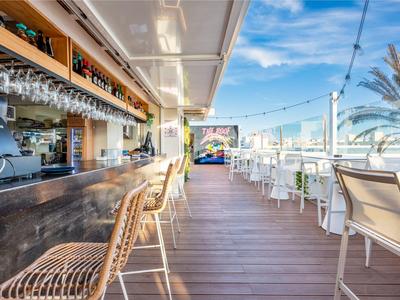 Offene Hotelbar mit Holzstühlen und Blick auf eine Terrasse mit Stühlen bei klarem Himmel.