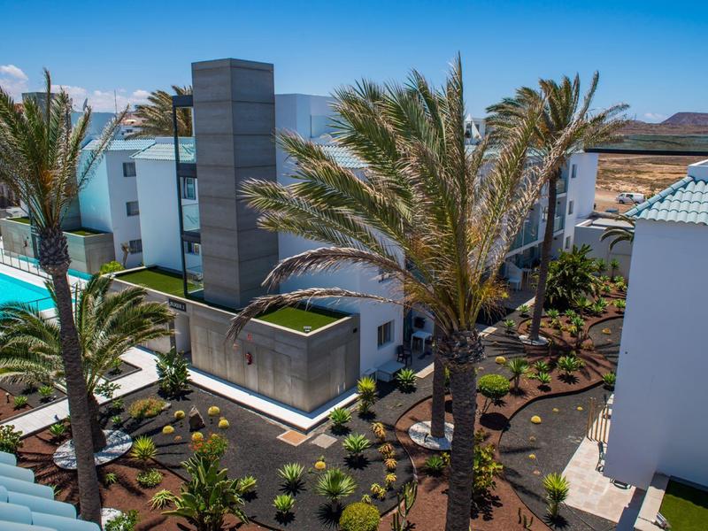 Modern hotel complex with pool, palm trees, and manicured gardens on a sunny day.