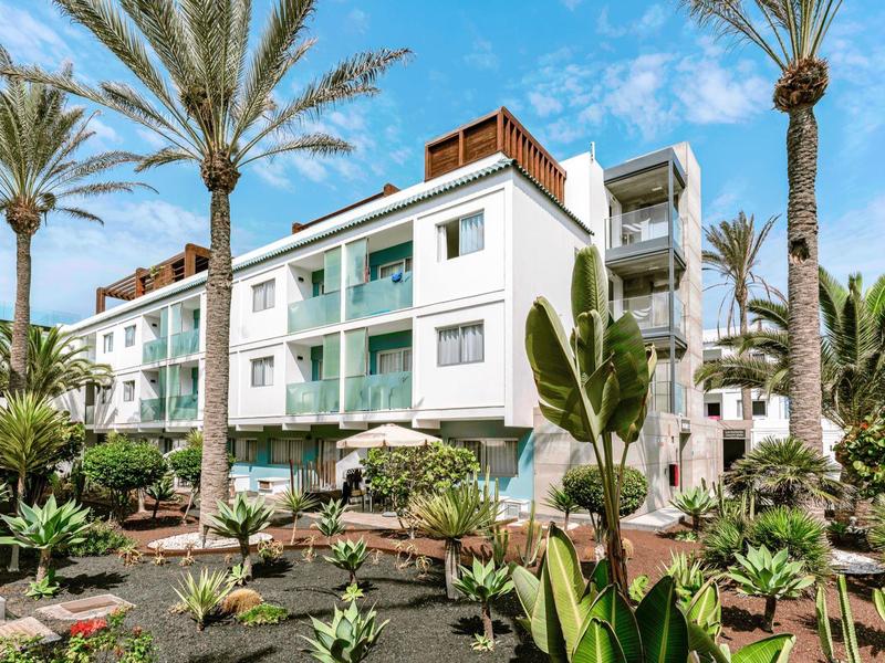 Three-story hotel with balconies surrounded by tropical plants and palm trees under a blue sky.