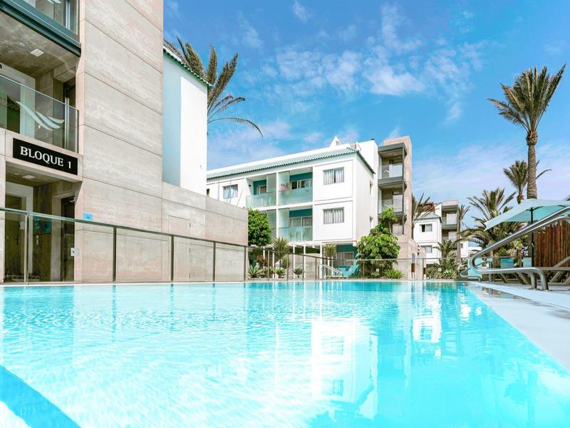 Modern hotel with a swimming pool, white buildings, and palm trees under a blue sky.