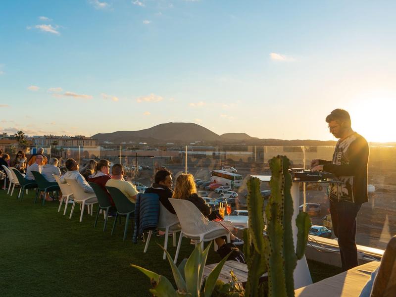 Guests sit outdoors at sunset with hill views and a waiter serving in an open-air setting.