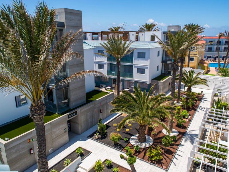 Modern hotel complex with white buildings, palm trees, and landscaped gardens under a blue sky.