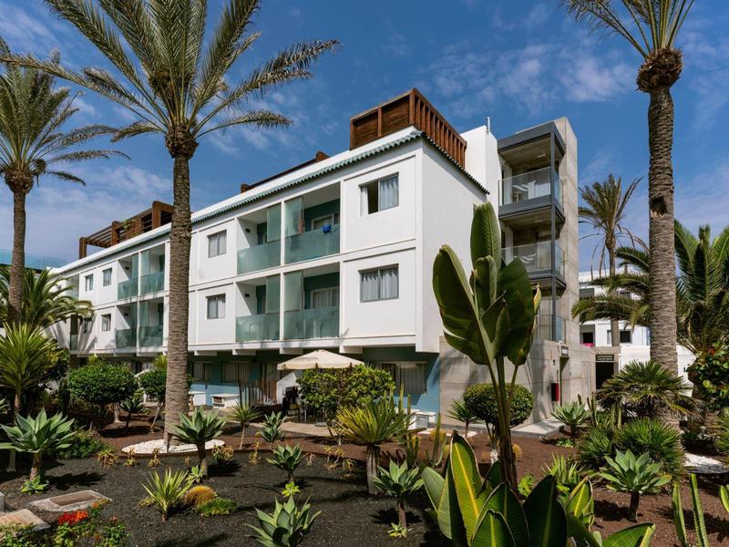 Modern white hotel building with balconies and tropical garden under blue sky.