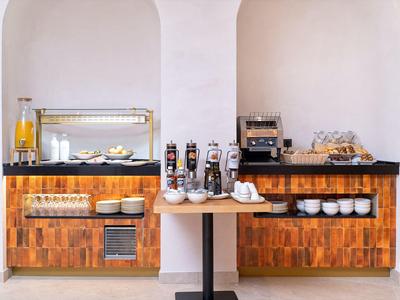 Area buffet con bevande, macchina del caffè e cibo caldo in una stanza luminosa.