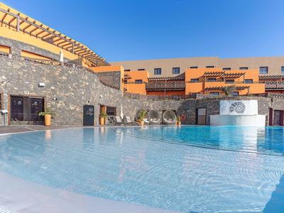 Moderner Außenpool mit Wasserfontäne und Sonnenterrasse in einer Hotelanlage unter blauem Himmel.