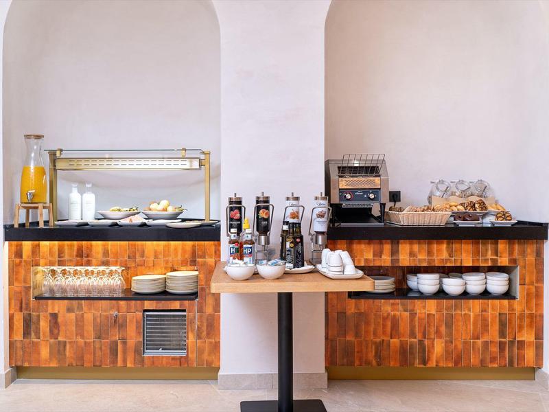 Area buffet con bevande, macchina del caffè e cibo caldo in una stanza luminosa.