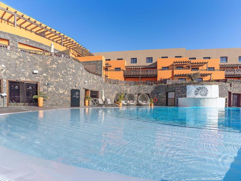 Piscina esterna moderna con fontana e terrazza soleggiata in un complesso alberghiero sotto un cielo azzurro.