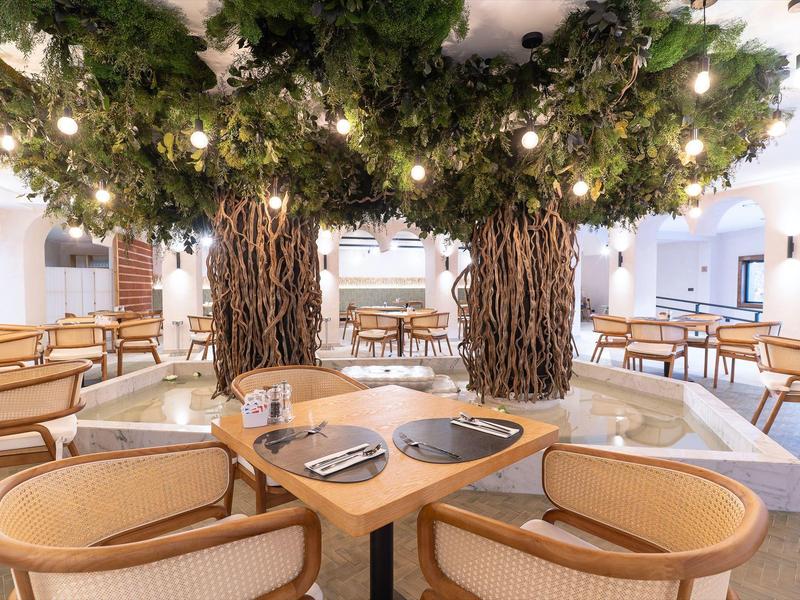 Area ristorante moderna con grandi alberi artificiali e tavoli apparecchiati.