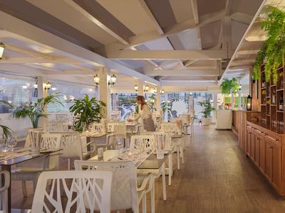 Restaurant lumineux avec mobilier blanc, sol en bois et grandes fenêtres donnant sur l'extérieur.