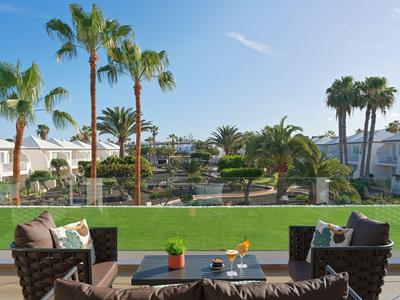 Vue depuis une terrasse d'hôtel sur un jardin avec des palmiers, des arbustes verts et des chaises longues.