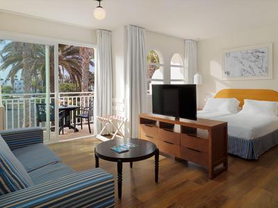 Chambre d'hôtel moderne avec balcon, lit double, canapé et télévision à écran plat sur sol en bois.