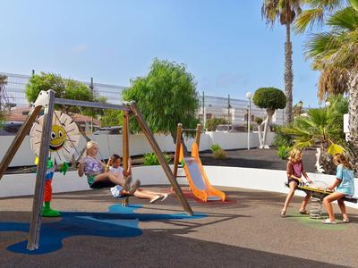 Des enfants jouent sur une aire de jeux avec balançoires et toboggan près des bâtiments d'un hôtel.