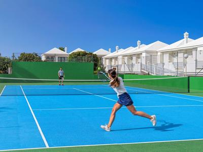 Femme jouant au tennis sur un court bleu avec des hôtels blancs en arrière-plan sous un ciel clair.