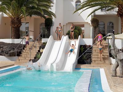 Des enfants glissent sur trois toboggans blancs à côté d'une piscine avec des palmiers et des statues d'éléphants.