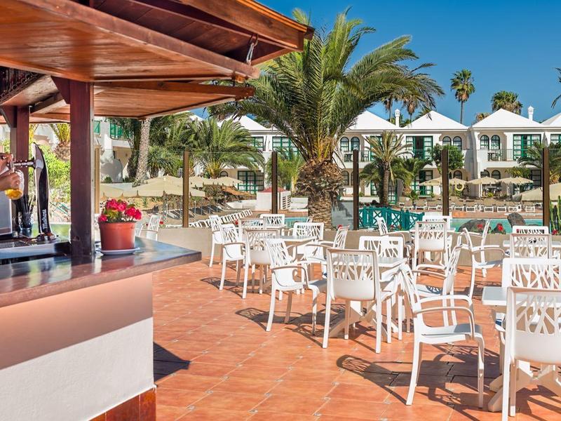 Outdoor bar with white chairs, tables, palm trees, and sun umbrellas in a sunny resort.