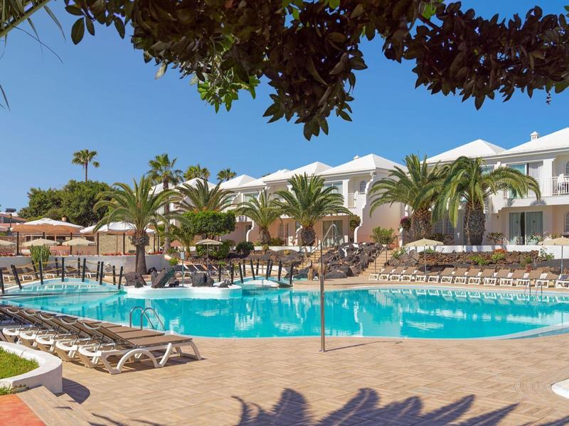 Outdoor hotel pool surrounded by sun loungers, umbrellas, palm trees, and white buildings under blue sky.