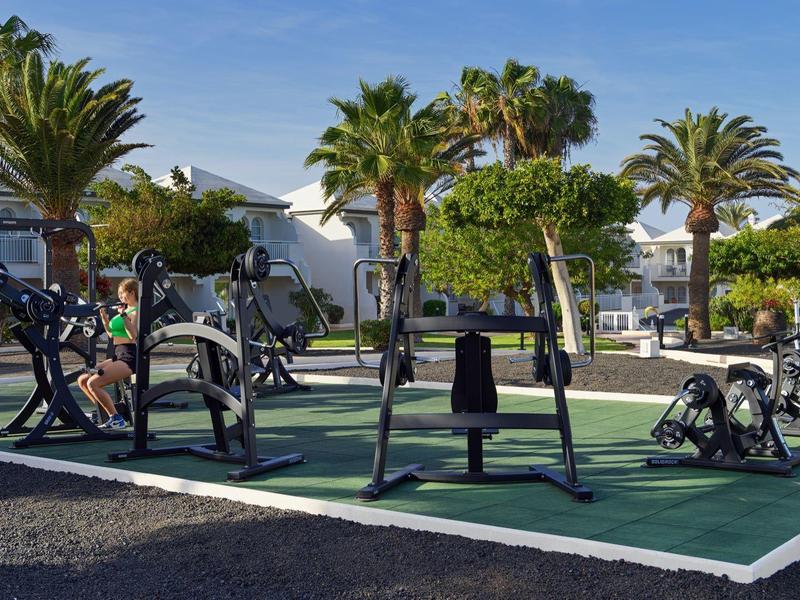 Outdoor gym area with exercise machines surrounded by palm trees under blue sky