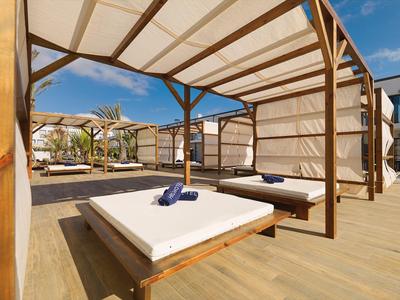 Outdoor wooden cabana with white mattress and towels under a clear blue sky at a resort.