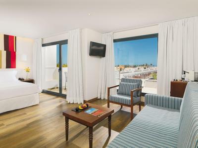 Bright hotel suite with wooden floor, king-size bed, sofa, and city view.