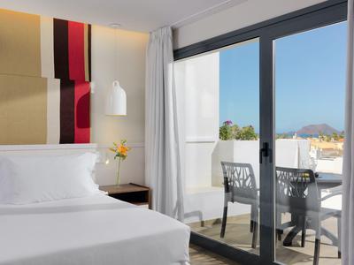 Bright hotel room with large bed, modern artwork, and balcony with outdoor furniture.