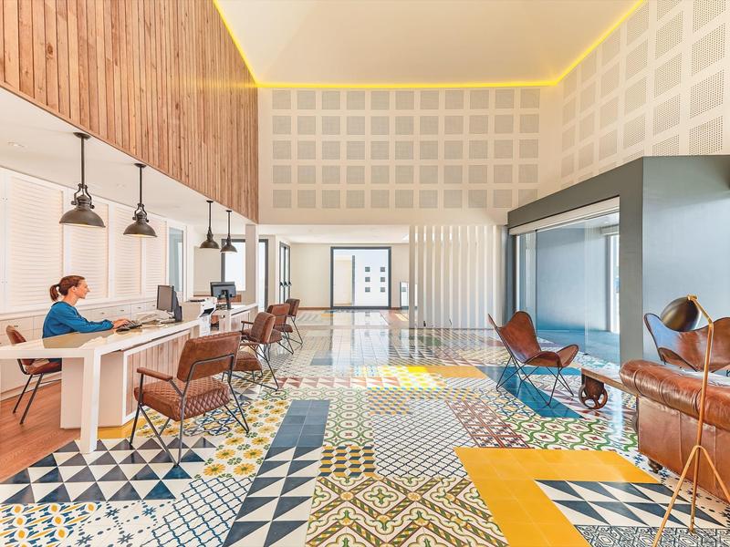 Bright hotel lobby with modern seating, patterned floor tiles, and wooden accents.
