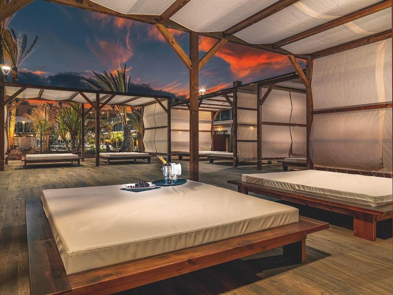 Outdoor cabanas with beds under a wooden pavilion at sunset.