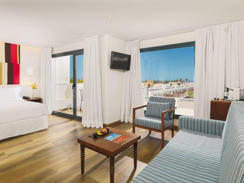 Bright hotel room with ocean view, modern seating, wooden floor, and adjacent bedroom.