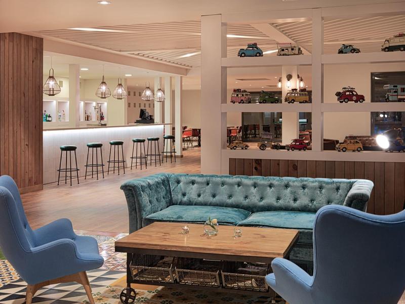 Modern hotel lounge with blue armchairs, teal sofa, wooden tables, and bar stools.