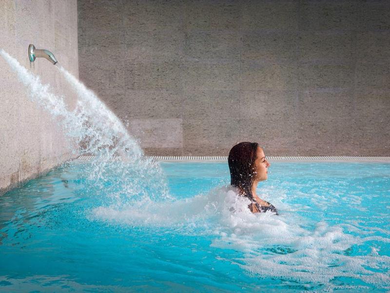 Person relaxing in an indoor pool with water flowing from two pipes into the water.