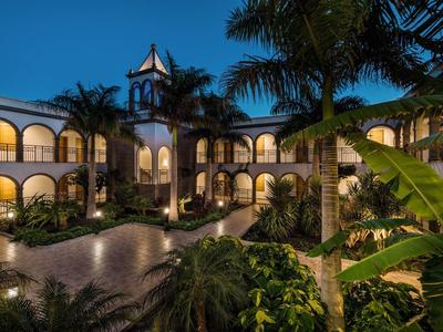 Vista serale di un hotel con balconi, camere illuminate e vegetazione tropicale.