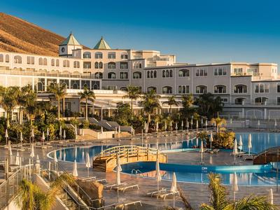 Hotel de lujo con gran piscina y palmeras bajo un cielo azul.