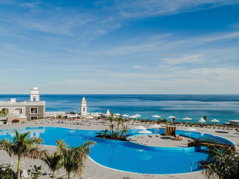 Grande piscina dell'hotel con palme, ombrelloni e vista sul mare sotto un cielo limpido