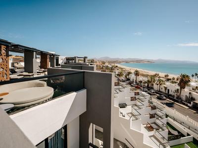 Balkon mit Sitzplatz und Blick auf moderne Gebäude und den Strand im Hintergrund unter blauem Himmel.
