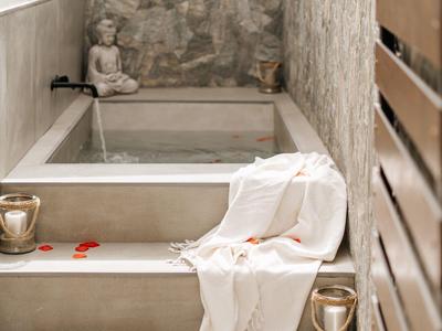 Ein heller Wellnessbereich mit Steinwand, Treppen, Badewanne, weißem Handtuch und roten Rosenblättern.