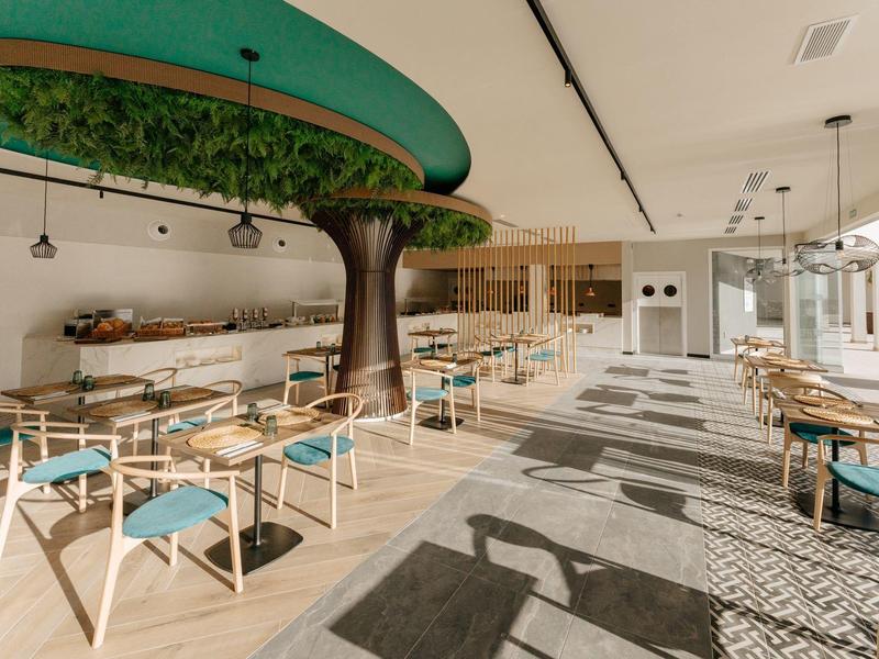 Sala da pranzo luminosa e moderna con tavoli in legno, sedie con cuscini blu e decorazione centrale ad albero.