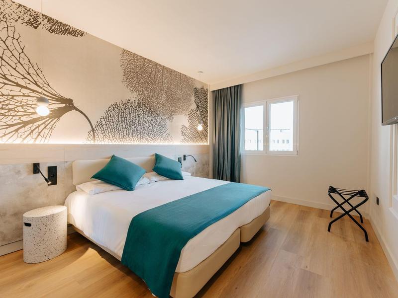 Camera d'hotel moderna con letto matrimoniale, cuscini turchesi e decorazioni, luminosa e minimalista.