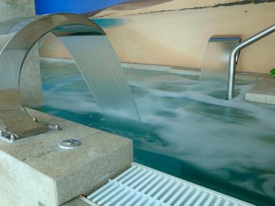 Piscina interna con cascata in acciaio inox e corrimano in metallo in una spa moderna.