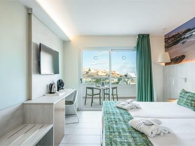 Camera d'hotel luminosa con letto matrimoniale, TV, scrivania e balcone con vista mare.